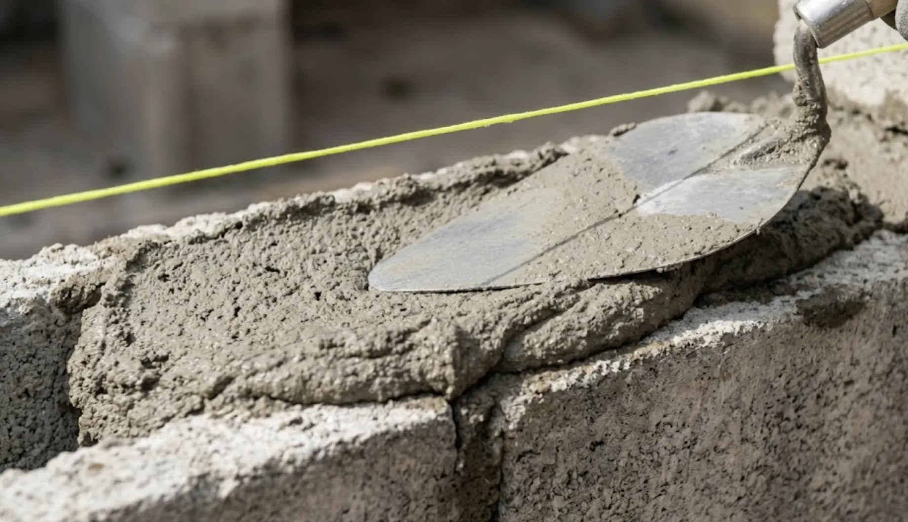 Gros plan sur une truelle étalant un lit de mortier épais de 2 à 3 cm sur le sommet irrégulier d'un vieux mur en parpaing, cordeau de maçon tendu au-dessus